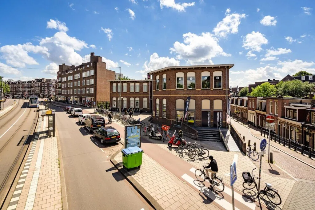 Straatbeeld van de Admiraal de Ruijterweg in Amsterdam met fietsers, auto’s, een tram en een karakteristiek bakstenen gebouw.